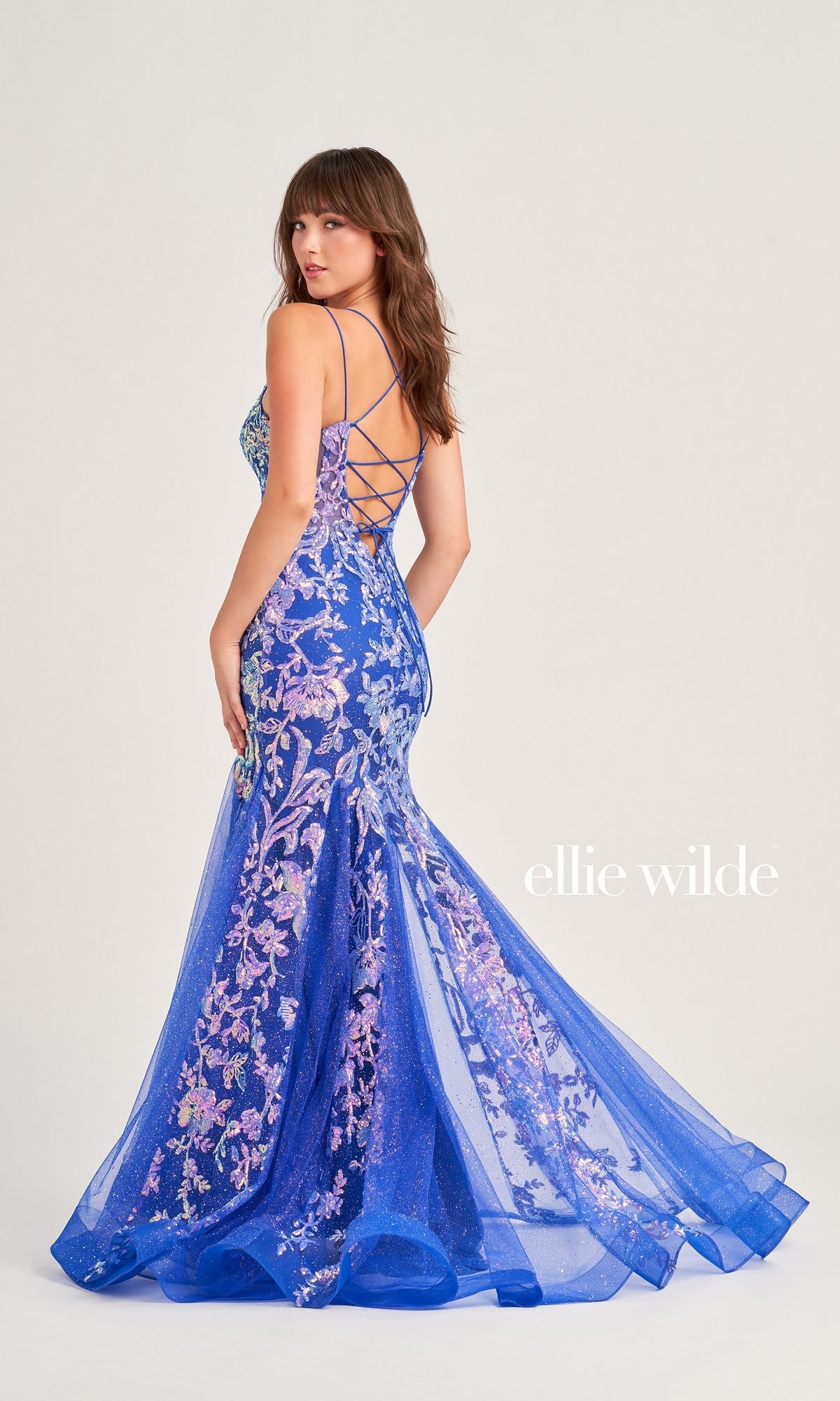 A woman in the Ellie Wilde Sequin Long Mermaid Prom Dress EW35008 stands facing away, glancing over her shoulder. The blue and purple floral gown features a lace-up back, sheer layers, and sparkling sequin details.